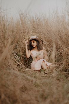A woman in a pink dress and straw hat sitting in a grassy field, exuding elegance.
