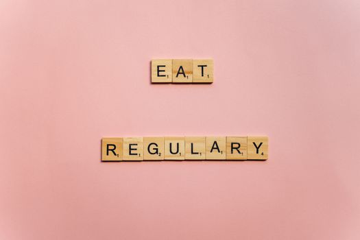 Wooden tiles spelling 'Eat Regularly' on a pink background, promoting healthy eating habits.