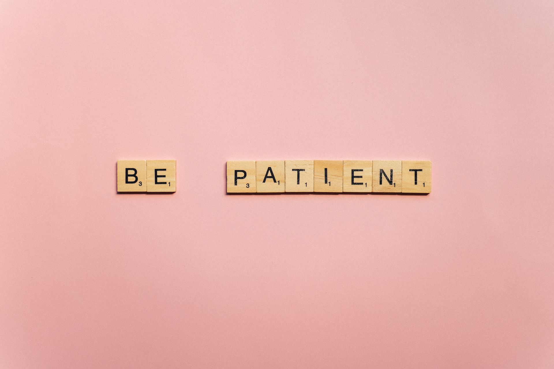 Inspirational words 'Be Patient' spelled with wooden tiles on a soft pink background.