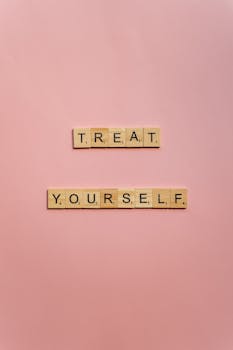 Wooden Scrabble tiles form the phrase 'Treat Yourself' on a soft pink background with ample copy space.