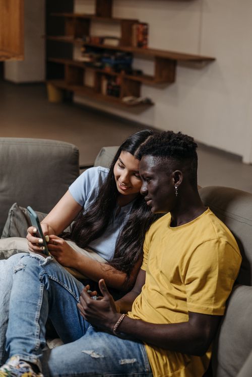 Free Young couple sitting together on a couch, sharing a moment looking at a smartphone indoors. Stock Photo