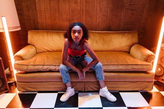 Young woman in a trendy outfit poses confidently on a vintage sofa with neon lights adding style.