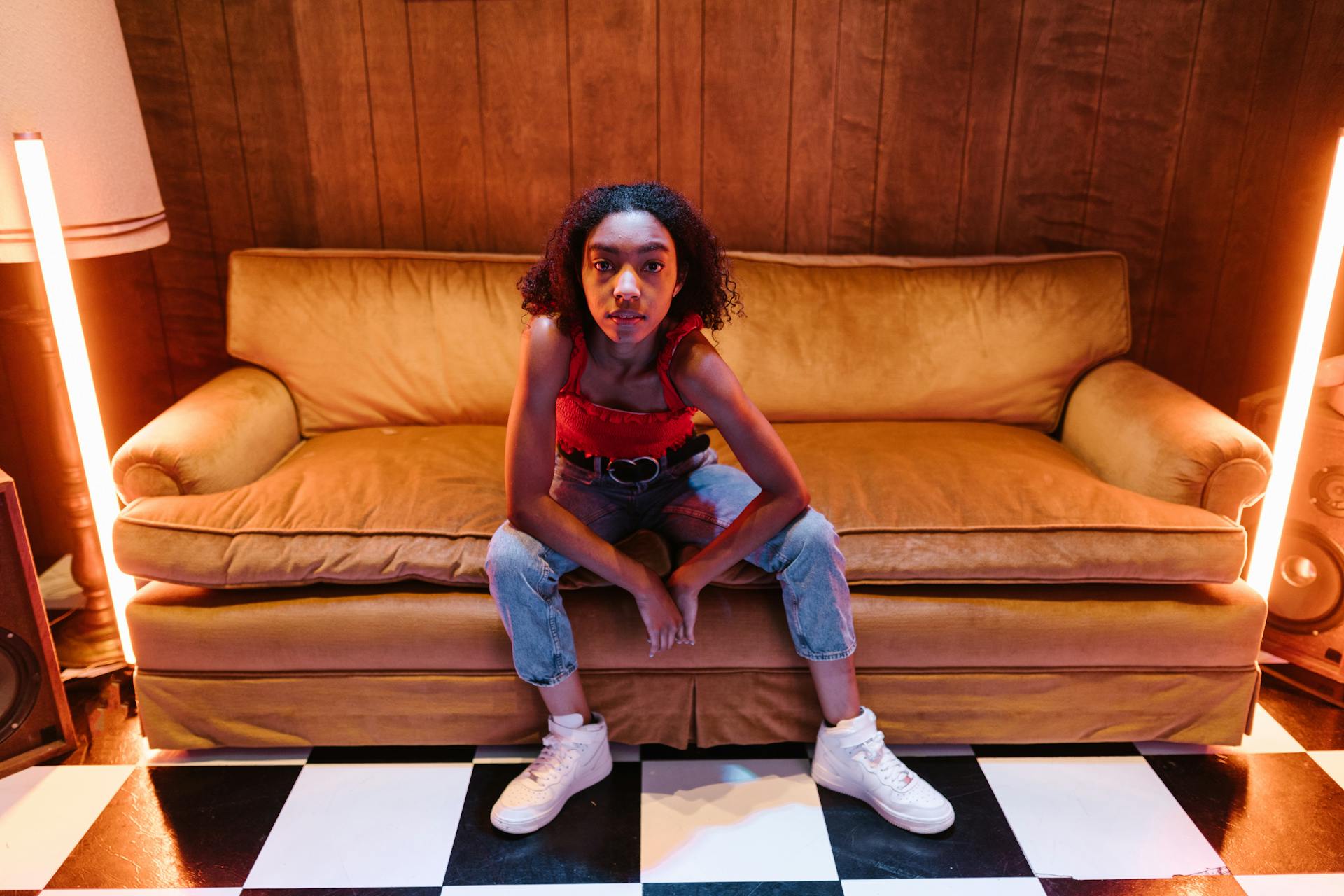 Young woman in a trendy outfit poses confidently on a vintage sofa with neon lights adding style.