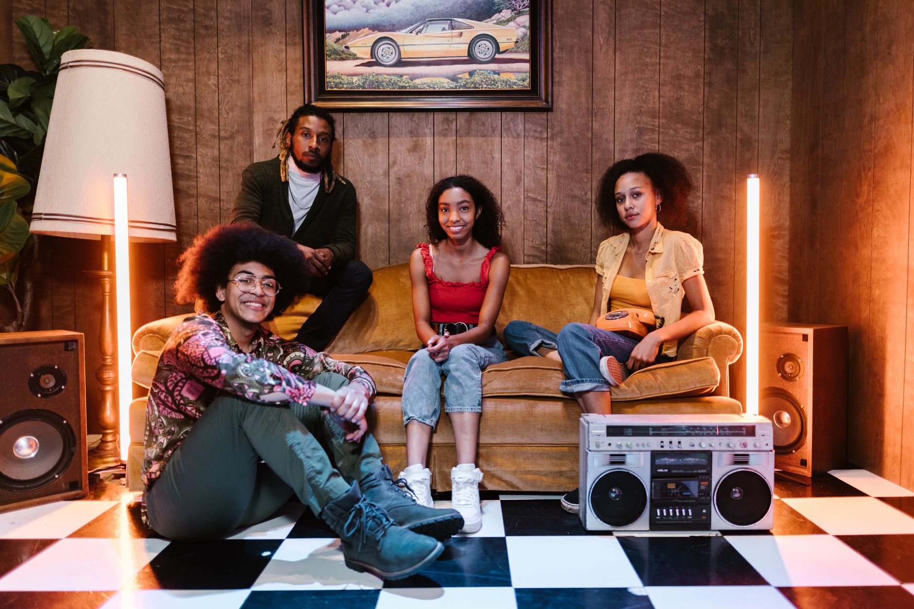 A diverse group of young adults sitting in a vibrant, retro-themed room with a boombox and vintage decor.