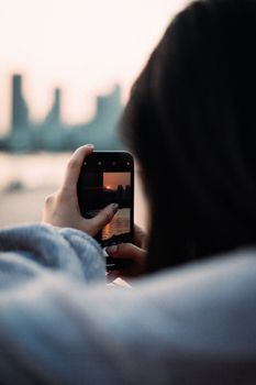 Woman photographing a sunset over a cityscape with a smartphone, showcasing urban beauty.