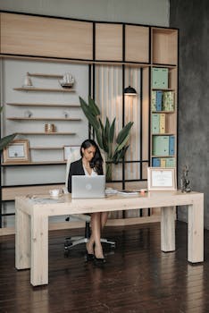 Woman in office working on a laptop at a modern wooden desk with shelves and decor.