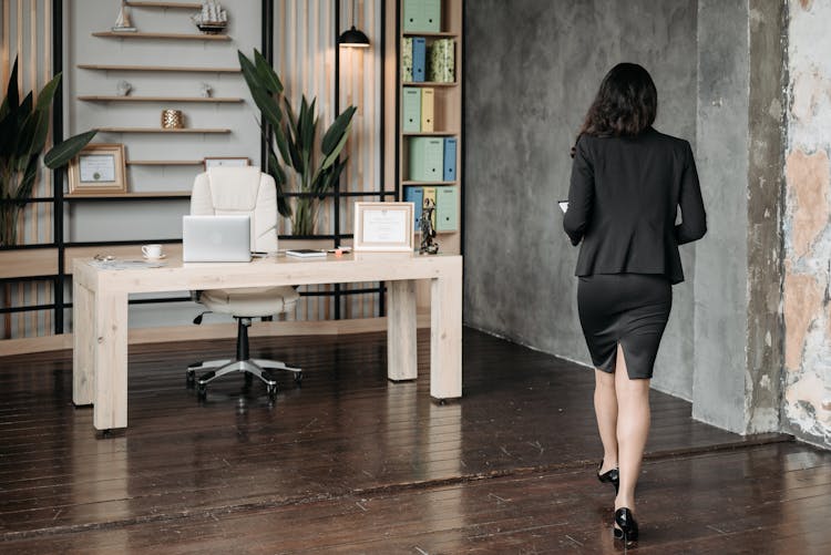 

A Woman In A Blazer And A Skirt Walking Towards Her Desk