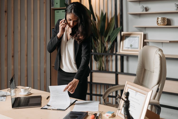 A Woman In Black Blazer Talking On The Phone While Looking At The Documents On The Table