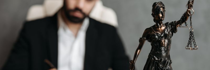 A lawyer in a suit writes at a desk with a Lady Justice statue in the foreground.