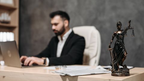 A businessman working on a laptop in an office with a Lady Justice statue in focus on the desk.