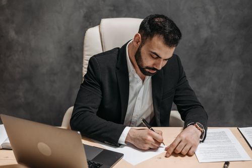 Free Businessman in black suit writing notes at desk with laptop and papers. Professional setting. Stock Photo