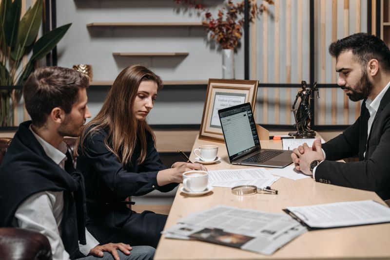 Two people seated across a table from a third, with an open laptop, printed pages, coffee cups and a small figure of Lady Justice between them.