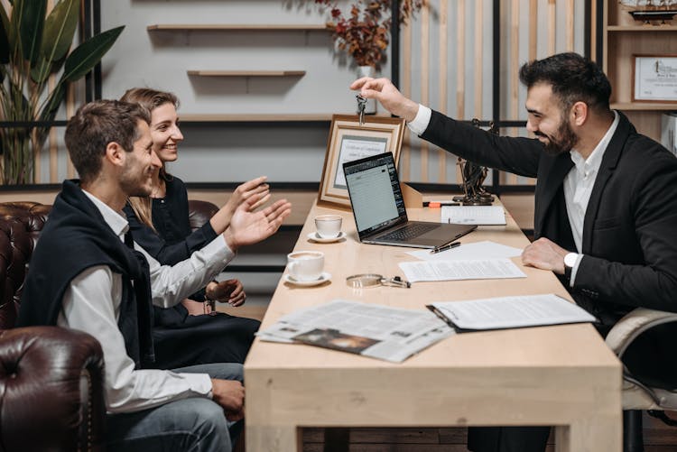 Smiling Employee Holding Keys Over Table Near Sitting Clients