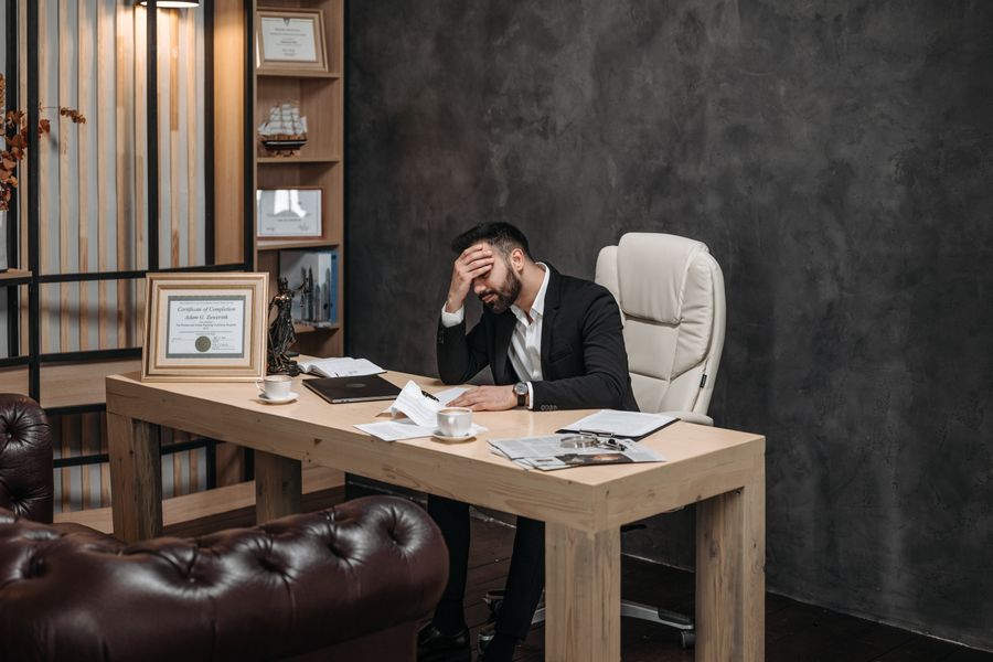 Businessman in a black suit looking stressed at an office desk, holding his head with papers scattered around.