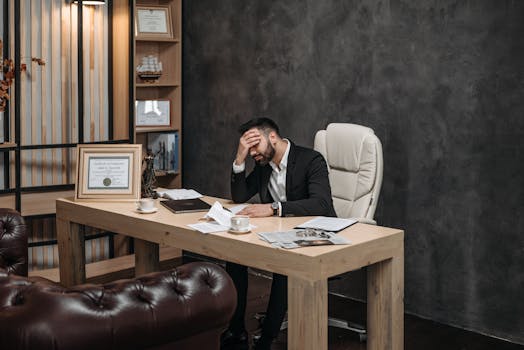 Businessman in a black suit looking stressed at an office desk, holding his head with papers scattered around.