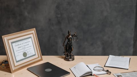 A lawyer's desk featuring a justice statue, diploma, laptop, magnifying glass, and documents.