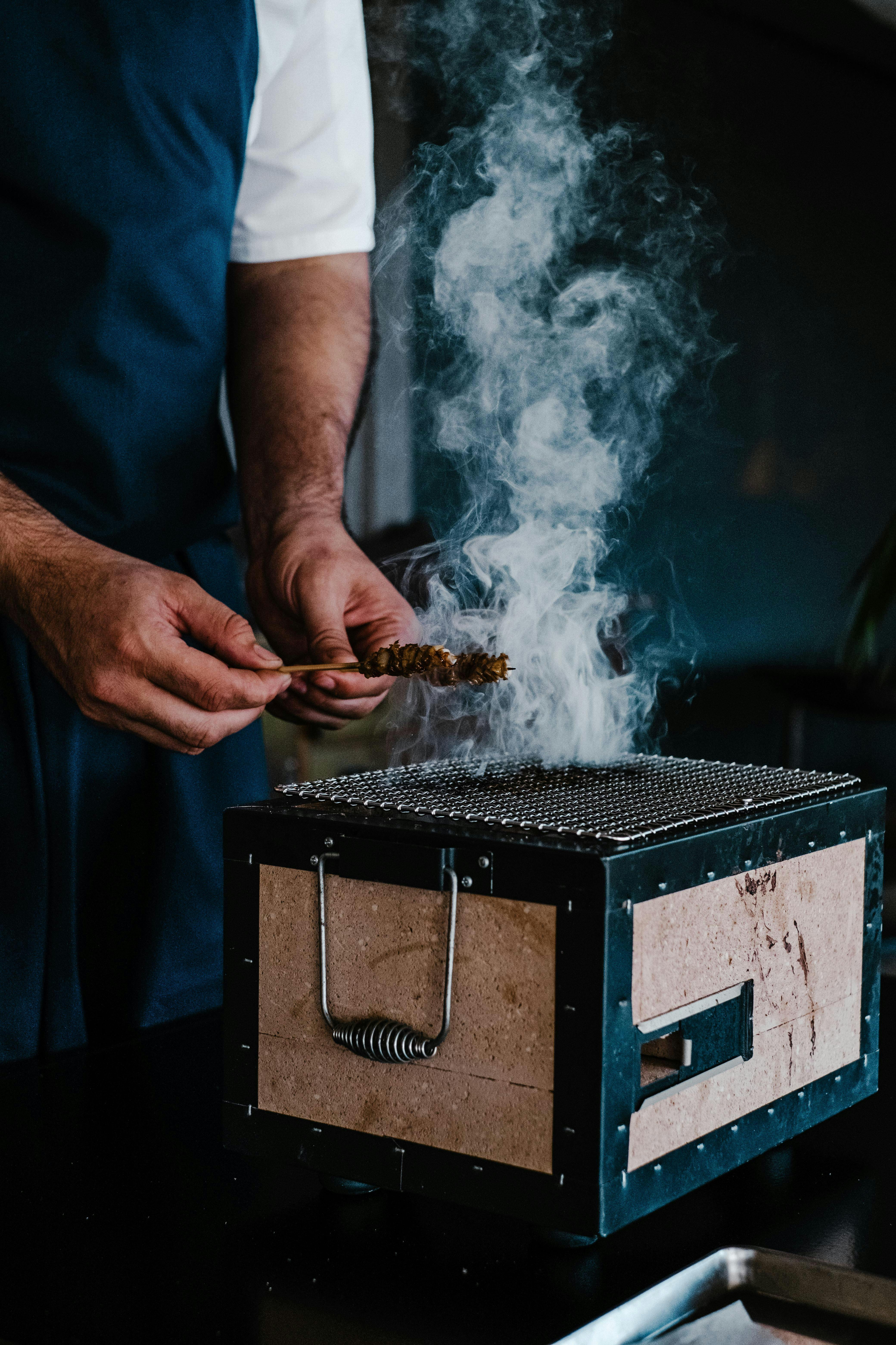 A chef expertly grills skewers on a hibachi, showcasing smoke and culinary skills indoors.