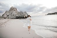 Woman in a white dress running along a tranquil beach with rocky formations at sunset.