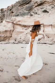 A woman in a white dress and hat walking on a sandy beach with rocky cliffs in the background.