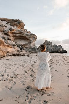 A woman in a white dress enjoys the picturesque rocky beach at sunset.