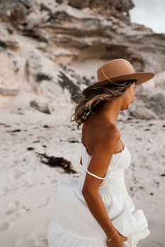 A carefree woman in a white dress and hat walks along a windy beach, embracing the moment.