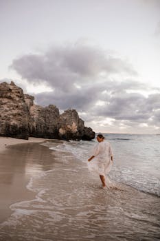 A lone woman walks along a tranquil beach at sunrise, surrounded by rocky cliffs under a cloudy sky.