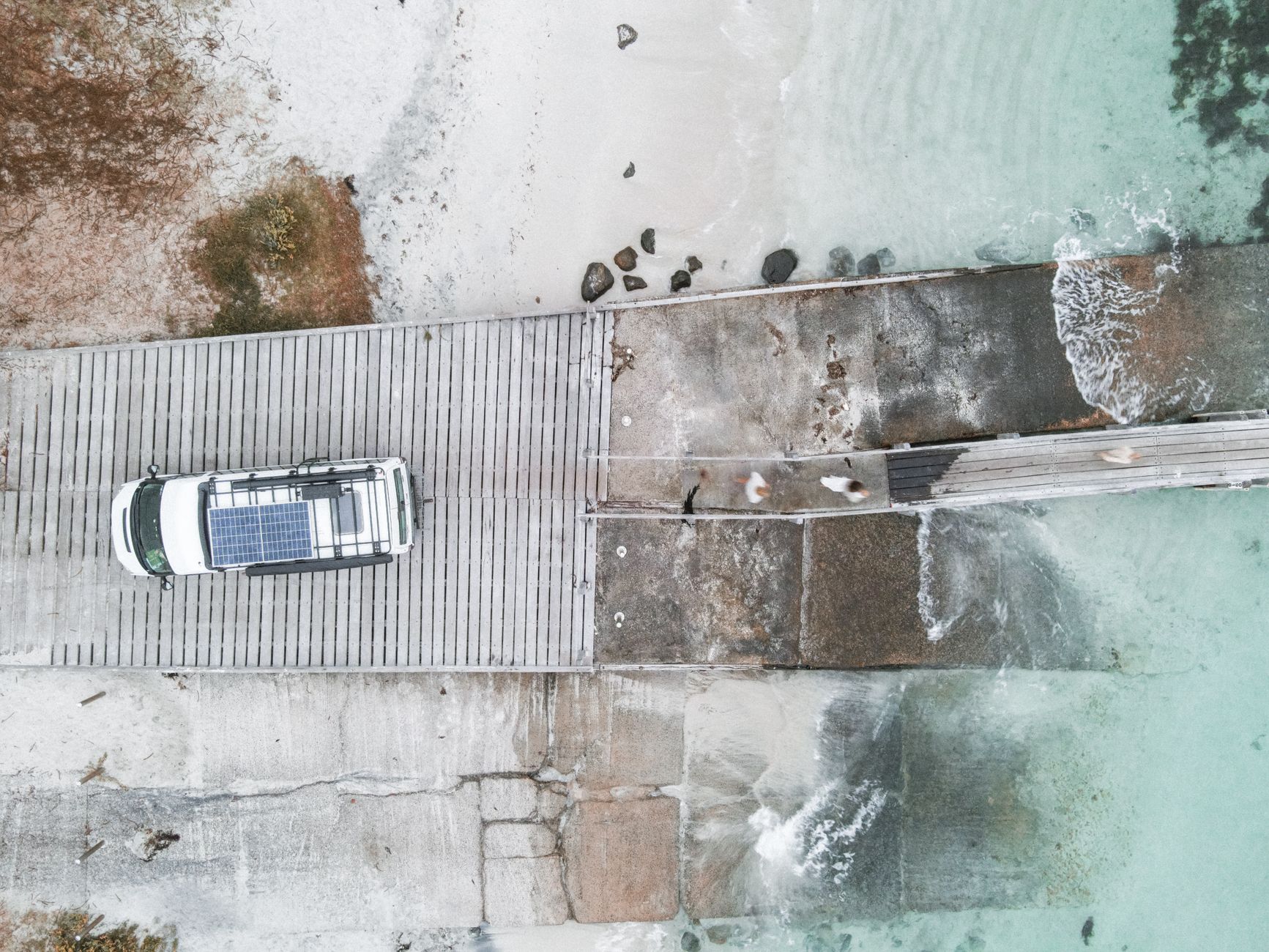 Aerial shot of a car with solar panels parked on a beachside pier by the turquoise sea.