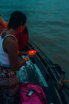 A woman holding a lit candle on a wooden boat during dusk on a peaceful river.