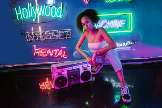 Fashionable woman posing with a boombox in a vibrant neon-lit setting, showcasing urban style.