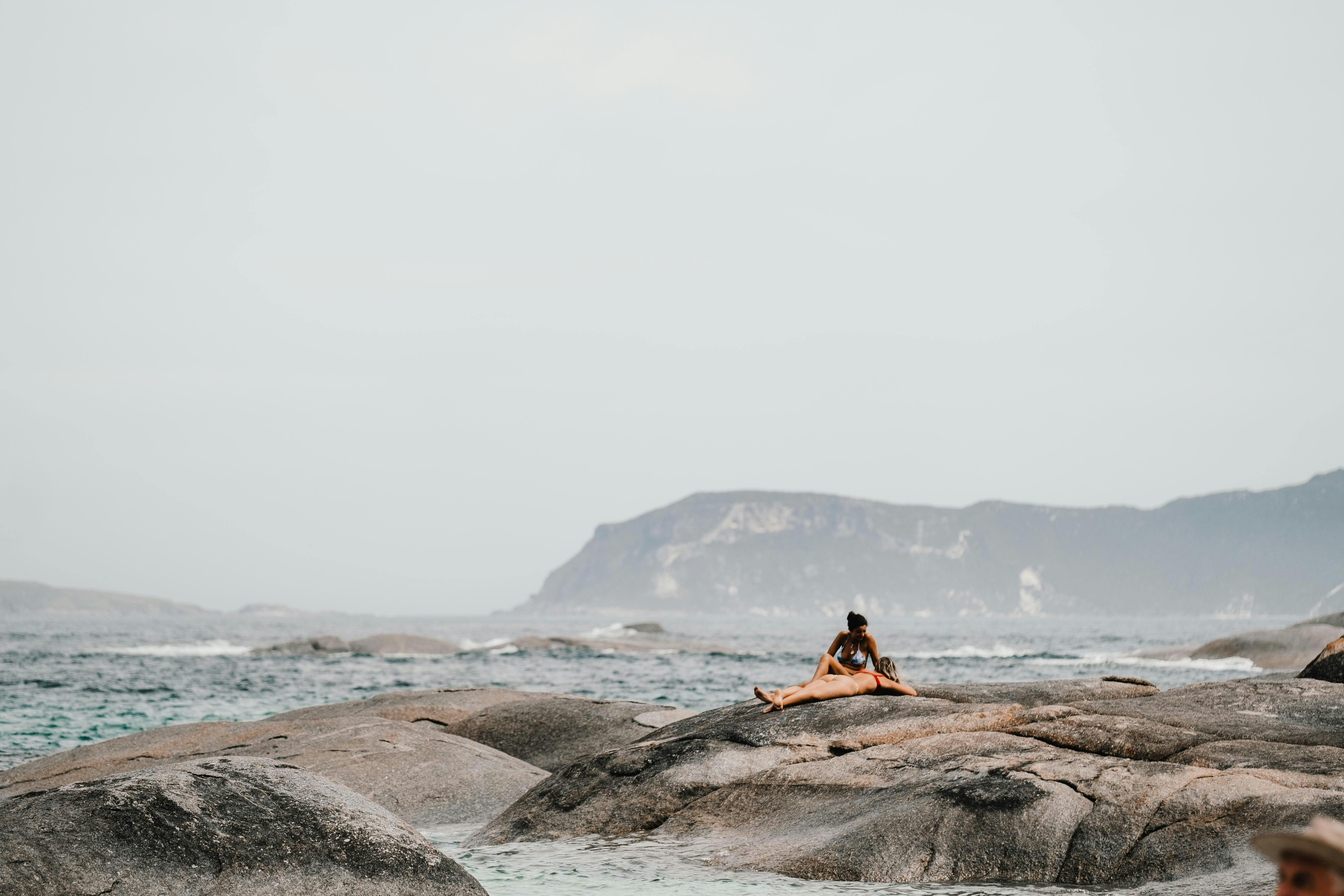 A woman sunbathing on rocky shore with ocean and mountains in the background.