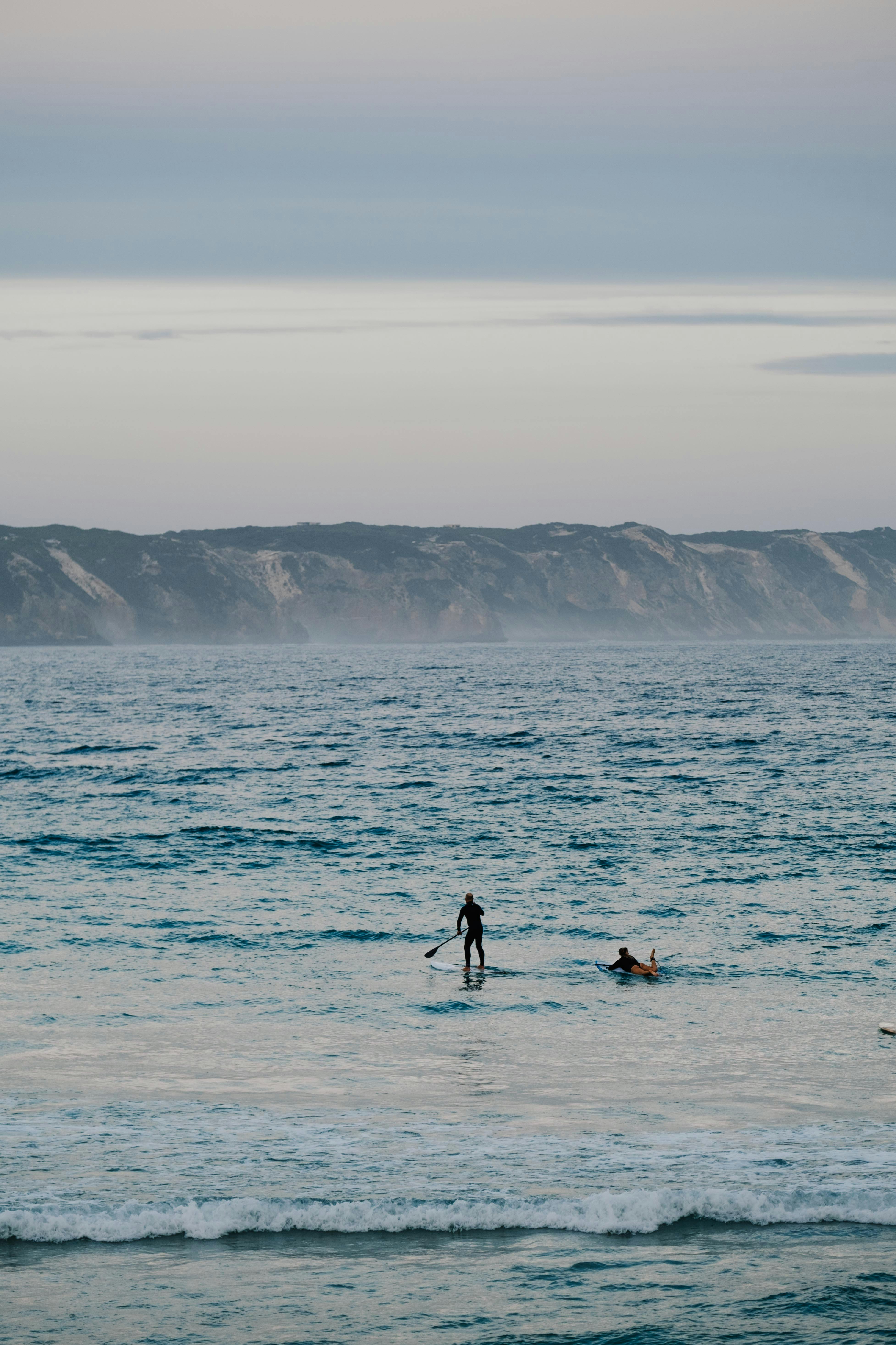 日落时分，一名孤独的桨板者在平静的水域中航行，柔和的天空下有远处的悬崖