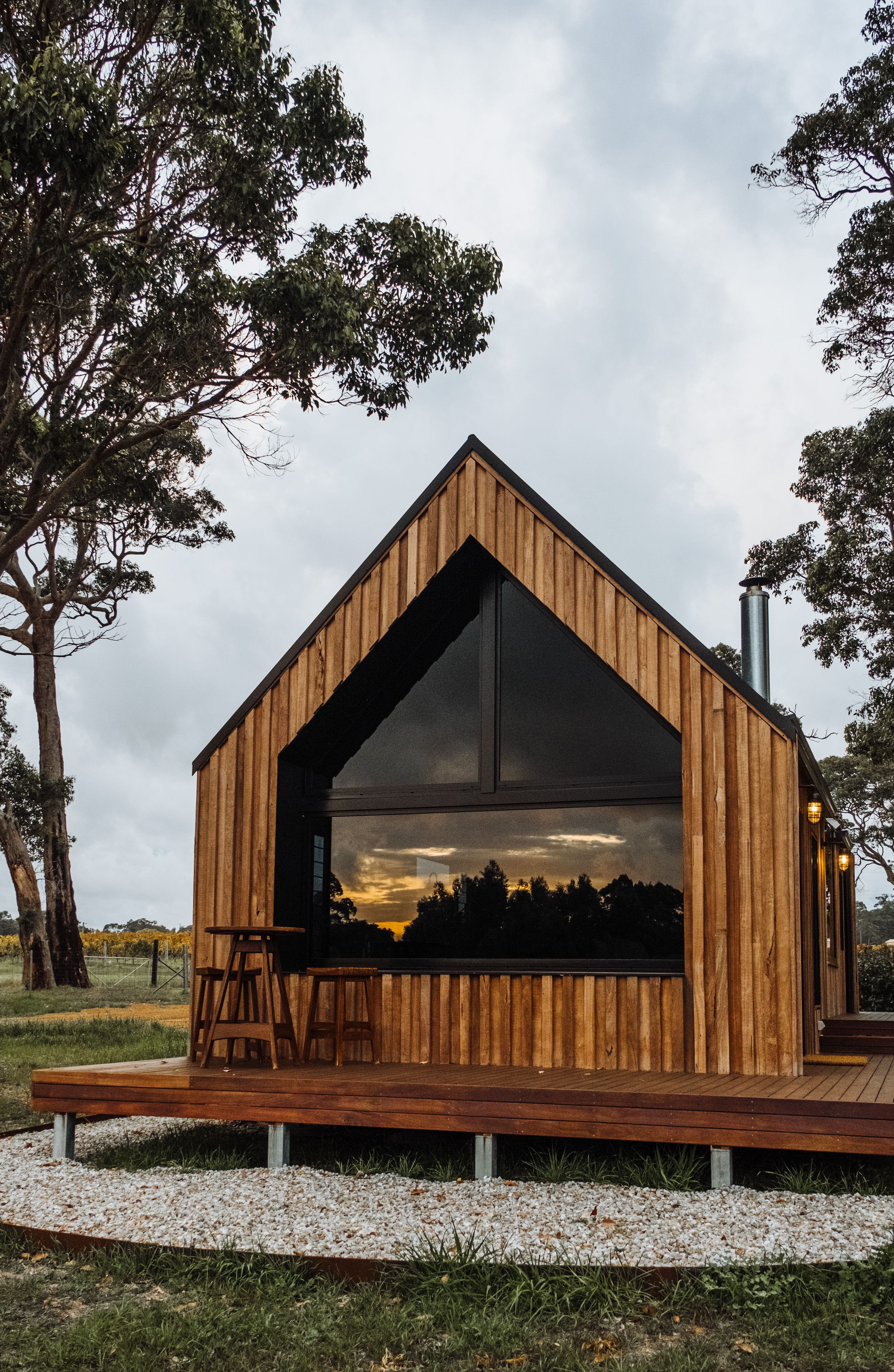 A cozy modern wooden cabin in a natural setting during sunset, with large windows reflecting the sky.