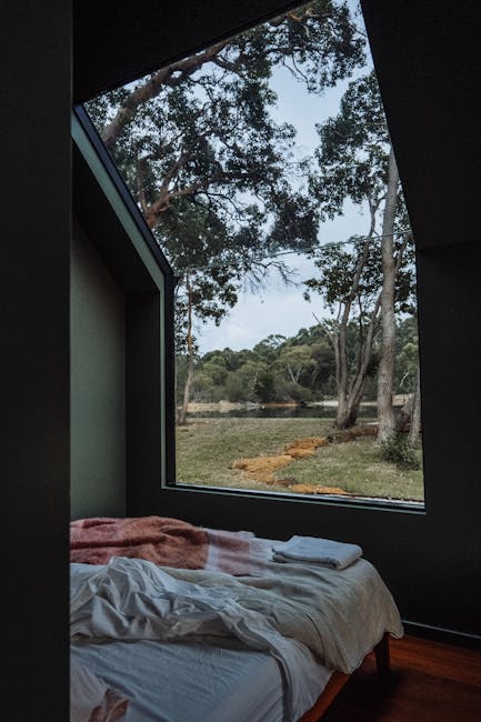 A cozy bed by a large window overlooking a serene forest landscape, perfect for relaxation.