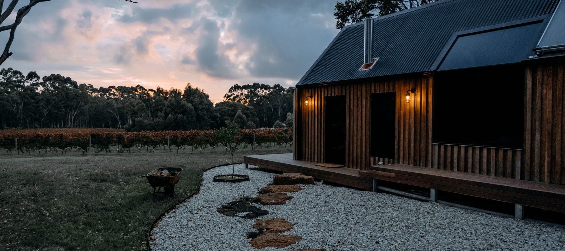 Studio de jardin en bois sombre au crépuscule, une allée de gravier mène à la porte