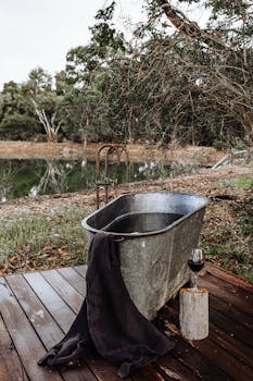 A rustic bathtub with a towel and glass of wine set by a peaceful lakeside.