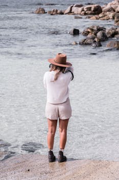 A woman in fashionable attire takes photos at a rocky seaside location.