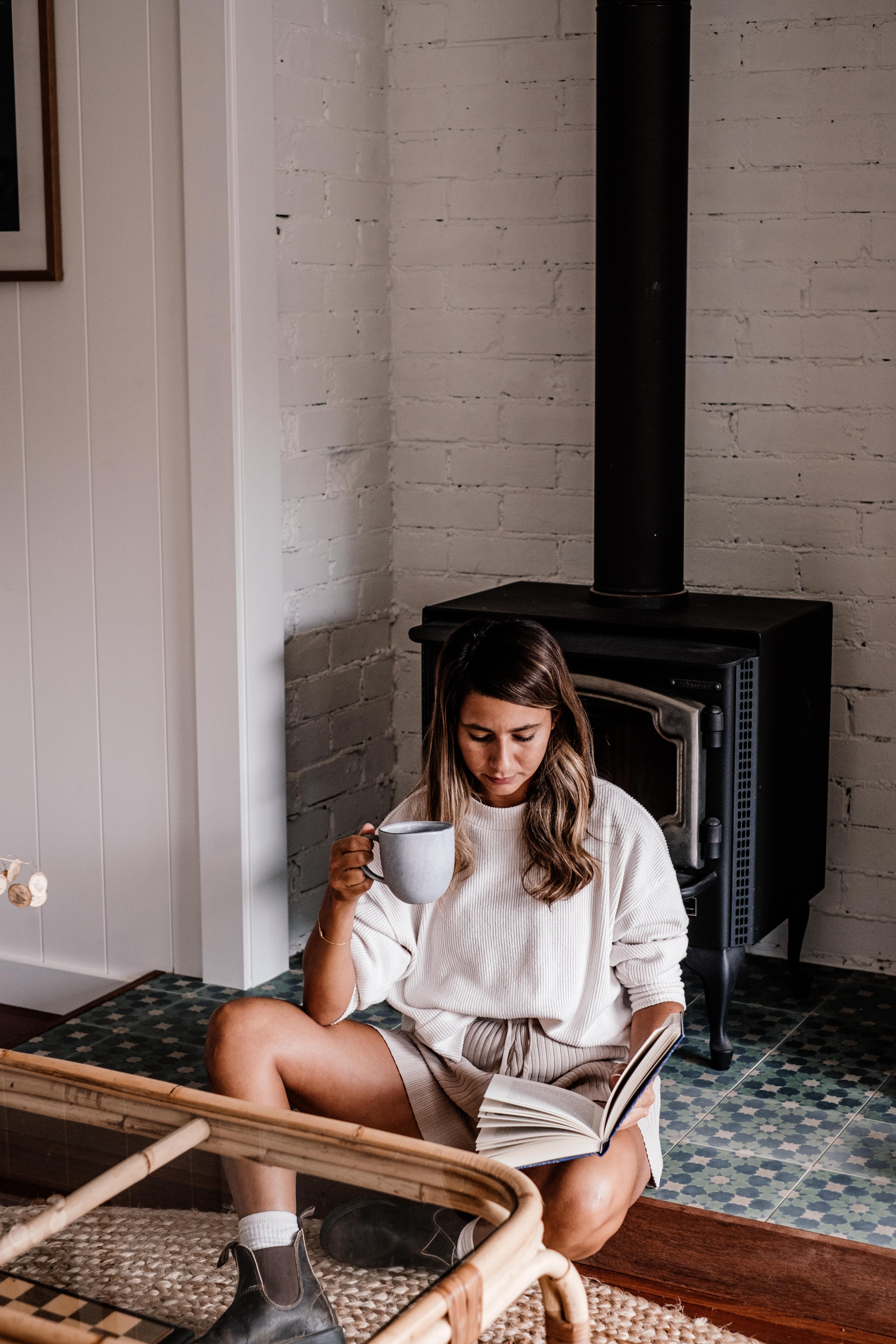 A woman enjoys a book with coffee in a cozy indoor setting with a fireplace.