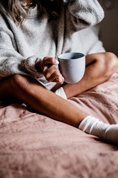 Relaxed morning scene with a person in a cozy gray sweater holding a coffee mug while sitting on a bed.