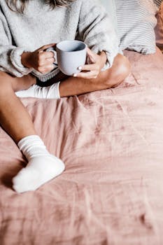 Person in warm sweater holding a mug while sitting on a bed, evoking a cozy morning vibe.