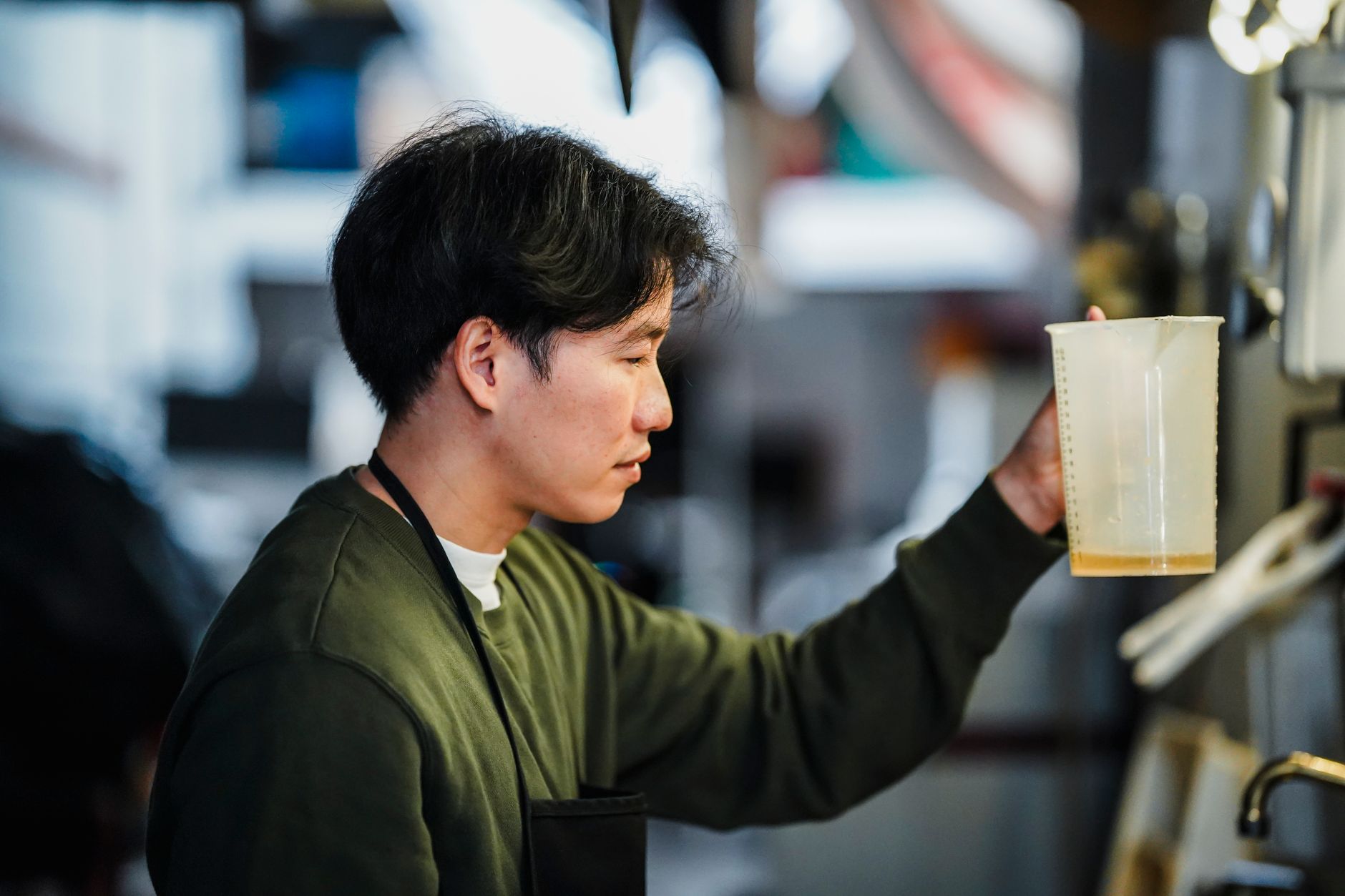 Asian man carefully holding a measuring cup with liquid, focused on precision in an indoor setting.