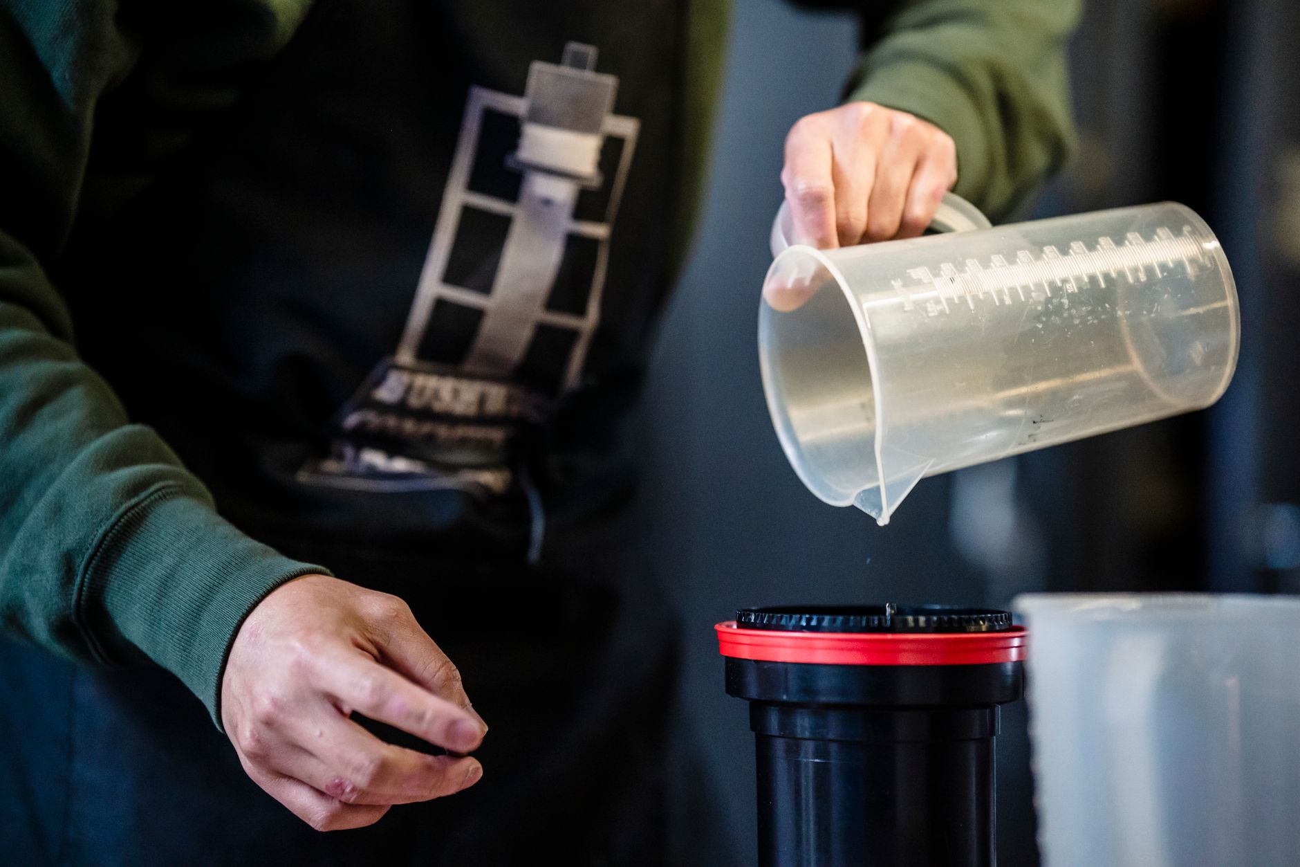 Hands pouring liquid from a measuring cup into a container, possibly for photo development.