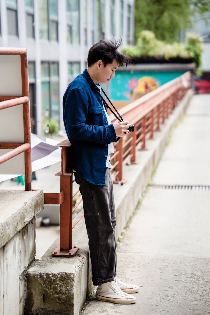 A young man in a denim jacket holding a camera, standing on an urban bridge.