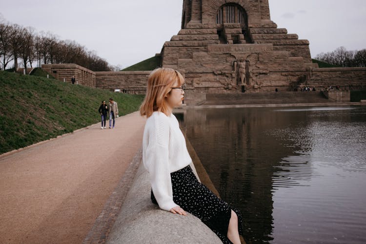 Young Woman Sitting On A Wall In Front Of The Monument To The Battle Of The Nations