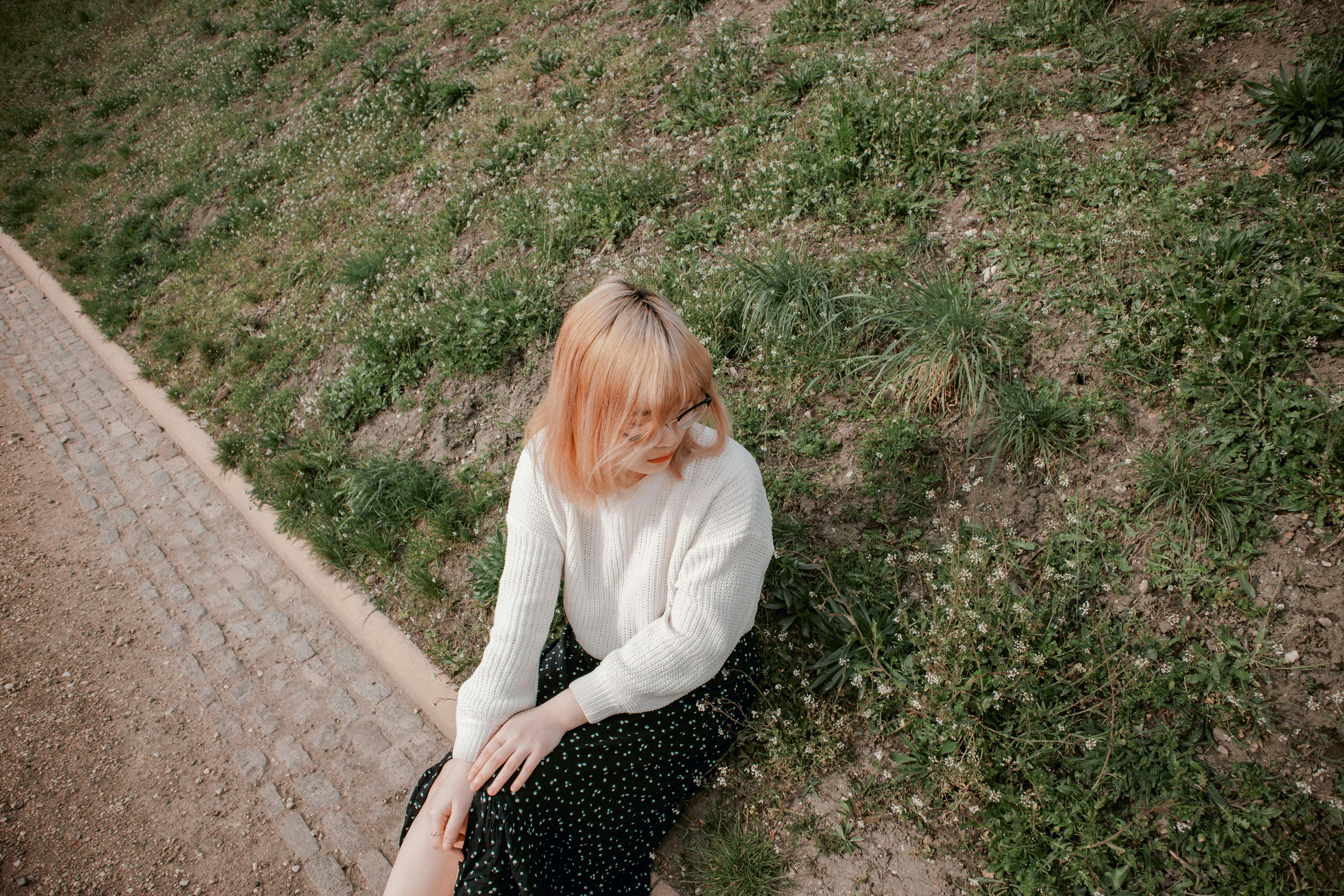 A young woman with blonde hair sits on a grassy slope, wearing a white sweater.