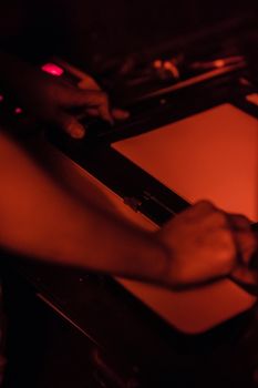 Hands working with photographic paper in a red-lit darkroom.