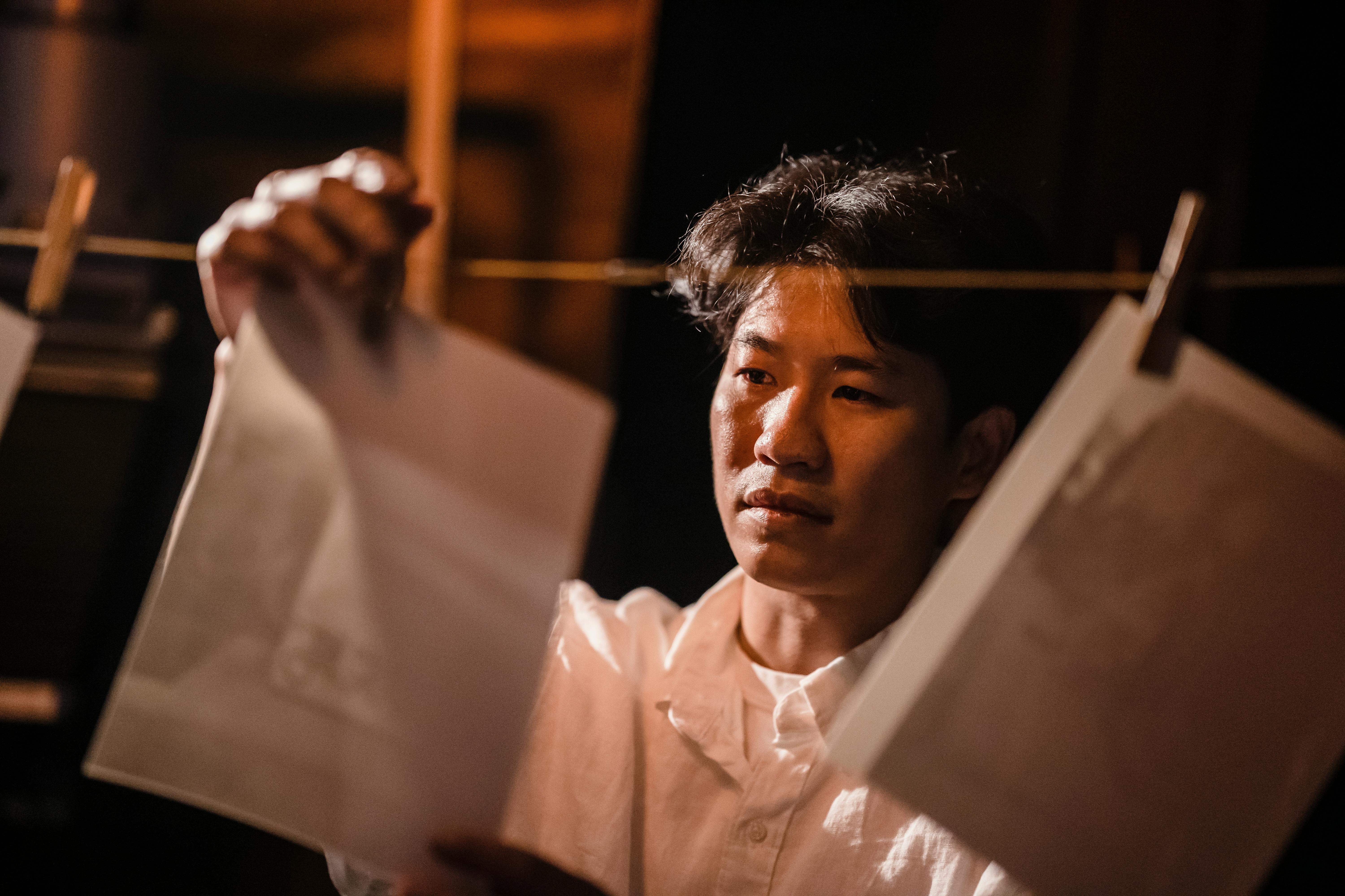 An Asian man carefully inspects printed photos in a darkroom setting, creating an artistic ambiance.
