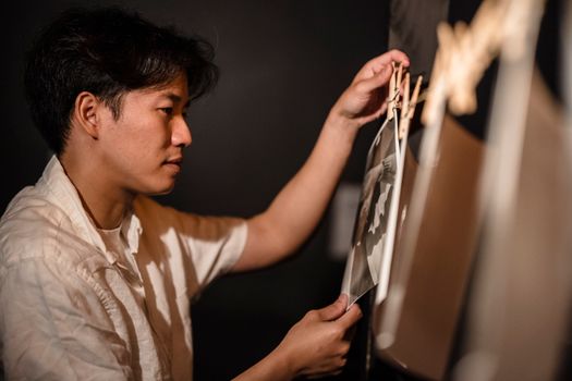 Asian man hanging photographs on a line in a darkroom setting.