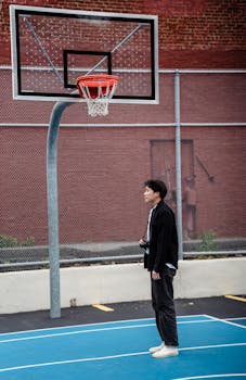 A man stands thoughtfully looking at a basketball hoop on an outdoor court.