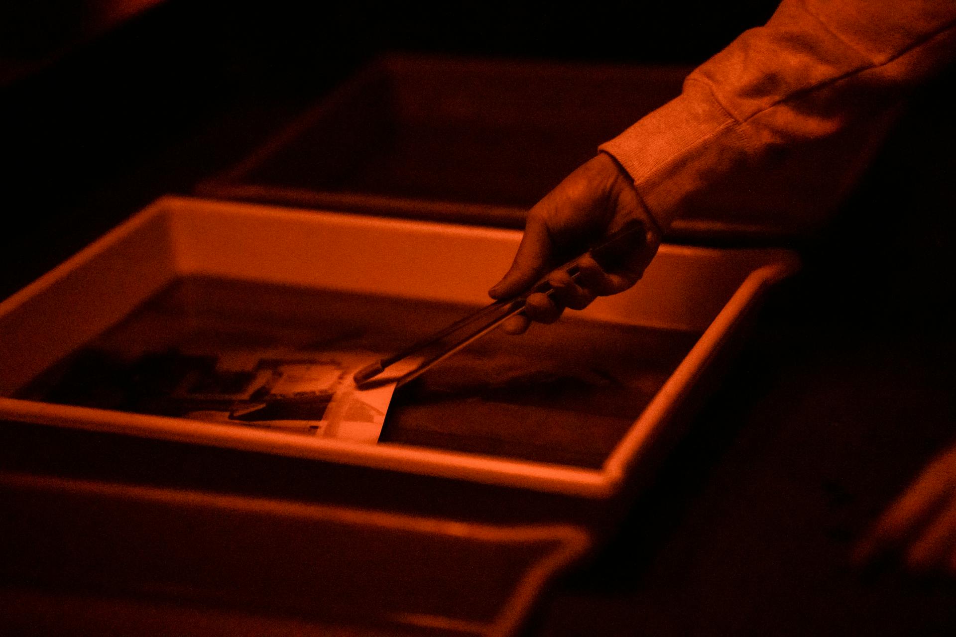 A detailed view of a hand using tongs to develop a photograph in a darkroom tray.