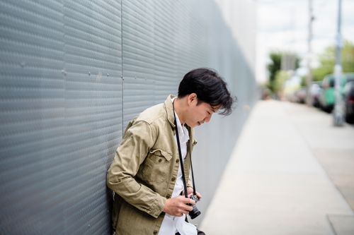 Free A young photographer leaning against a textured urban wall, looking down at a camera. Stock Photo
